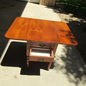 May include: A wooden drop-leaf table with a warm brown finish, featuring two drawers with dark metal pulls. The table has a rectangular top with rounded corners and turned legs. One drawer is partially open, revealing a white interior lining.