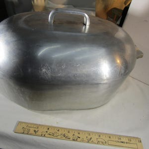 May include: A large, oval-shaped silver-coloured aluminium roasting pan with a matching lid and side handles. A wooden ruler is placed in front of the pan, showing a length of approximately 30.5 cm for the base of the roaster.