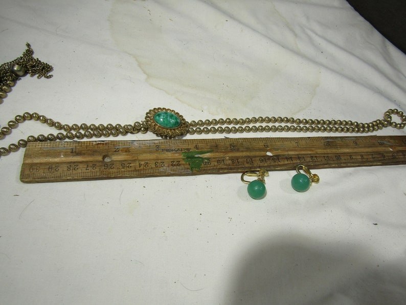 Peut inclure: Un collier en or avec un pendentif en pierre verte et des boucles d'oreilles assorties en pierre verte. Le collier est compos&eacute; de petites perles dor&eacute;es et le pendentif est une pierre verte ronde sertie dans un cadre en or. Les boucles d'oreilles sont &eacute;galement des pierres vertes rondes serties dans des cadres en or.