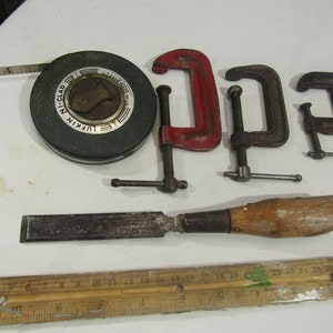Op de afbeelding: Een verzameling vintage gereedschap, waaronder een groene rolmaat met de tekst "LUFKIN NI-CLAD", drie C-klemmen, waarvan één met rood geverfde handvatten, en een beitel met een houten handvat.