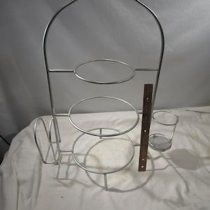 May include: Silver metal tiered serving stand with three round shelves and a side rack for napkins. The stand has a decorative scroll design at the top.