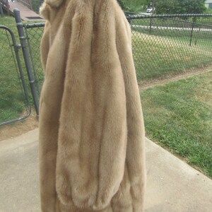 May include: A long, brown faux fur coat with a high collar. The coat is hanging on a white stand against a backdrop of green grass and a chain link fence.