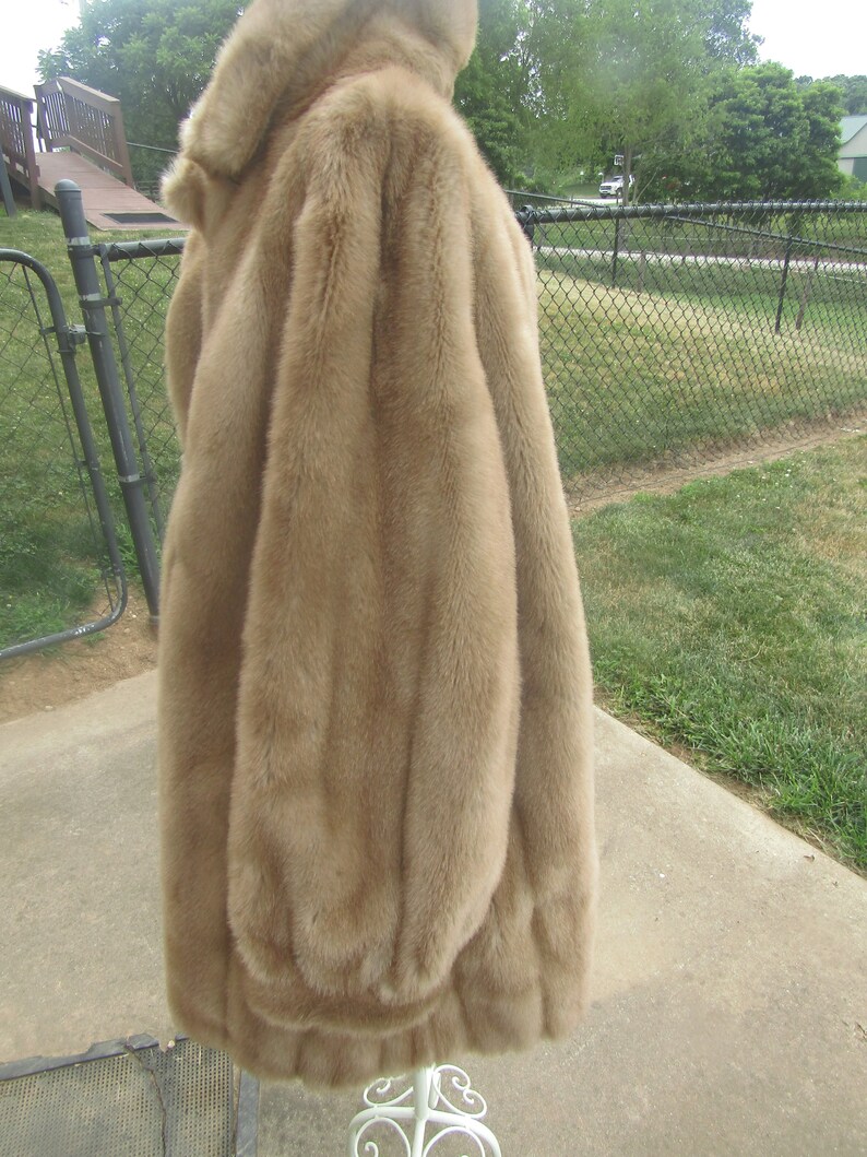 May include: A long, brown faux fur coat with a high collar. The coat is hanging on a white stand against a backdrop of green grass and a chain link fence.