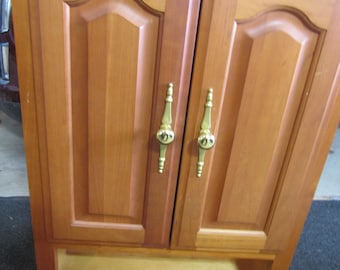 May include: A wooden cabinet with two doors and a shelf. The doors have ornate brass handles. The cabinet is a light brown color.