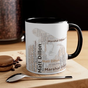 May include: A white ceramic mug with a black rim and handle. The mug features a word cloud design with the words "Matt Dillon" and "Marshal Dillon" repeated throughout. The design is in a brown color and is shaped like a person.