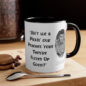 Könnte beinhalten: Eine weiße Keramiktasse mit schwarzem Rand und Henkel. Die Tasse zeigt ein Schwarzweißbild eines Mannes mit einem Zitat, das lautet "Ain't we a Pickin' OUR Peaches 'Fore They're Fuzzed Up GOOD?" Das Zitat wird "Sheriff Andy Taylor The Andy Griffith Show" zugeschrieben.