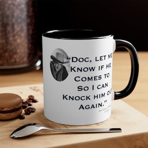 May include: White ceramic mug with a black rim and handle. The mug features a black and white photo of a man wearing a cowboy hat and a quote that reads "Doc, let me know if he comes to so I can knock him out again."