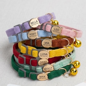 May include: A selection of cat collars in various colours, including purple, blue, pink, brown, yellow, red, and green. Each collar features a gold name tag with a cat's name and phone number. Gold bells are attached to the collars.
