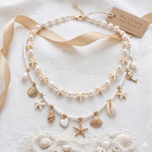 May include: Double-strand white beaded necklace with gold accents and seashell charms. The necklace features starfish, pearls, and a tag that reads "SEA CHARM NECKLACE HANDMADE". Displayed on a white surface with sand and seashells.
