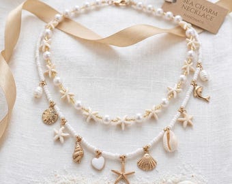 Könnte beinhalten: Zweisträngige, weiß-perlenbesetzte Halskette mit goldenen Akzenten und Muschel-Anhängern. Die Halskette hat Seesterne, Perlen und ein Etikett mit der Aufschrift "SEA CHARM NECKLACE HANDMADE". Präsentiert auf einer weißen Oberfläche mit Sand und Muscheln.