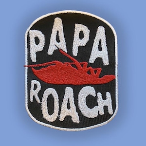 Puede incluir: Parche bordado con las palabras "PAPA ROACH" en blanco, arqueadas alrededor de un diseño de insecto rojo, sobre un fondo negro. El parche tiene un borde blanco y está sobre un fondo azul claro.