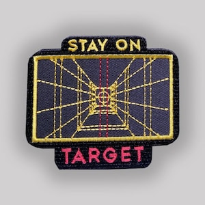 Könnte beinhalten: Ein gestickter Aufnäher in Schwarz und Gold mit dem Text "STAY ON TARGET" und einem stilisierten Bild eines Zielsystems.