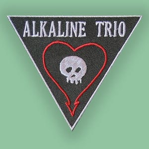 Puede incluir: Parche triangular con el texto "ALKALINE TRIO" en blanco. Presenta un corazón rojo con una calavera blanca en el centro y una flecha hacia abajo. El parche está sobre un fondo gris oscuro.