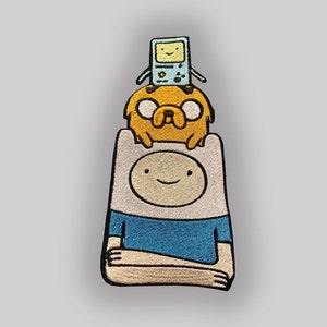 Può includere: Una toppa ricamata con Finn l'Umano della serie animata Adventure Time. Finn indossa una camicia blu e ha una testa bianca con un viso sorridente. Jake il Cane è seduto sulla testa di Finn, e un dispositivo simile a un Game Boy è sulla testa di Jake.