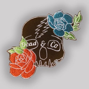 Può includere: Una toppa ricamata con un teschio nero con un design a forma di fulmine e il testo "Dead & Co" in bianco. Il teschio è adornato con tre fiori: una rosa rossa, una rosa blu e una foglia verde.