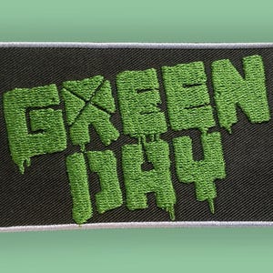 Puede incluir: Parche bordado con el nombre de la banda "GREEN DAY" en letras verdes y goteantes sobre un fondo rectangular negro. El parche tiene un borde blanco, adecuado para aplicar en ropa o accesorios.