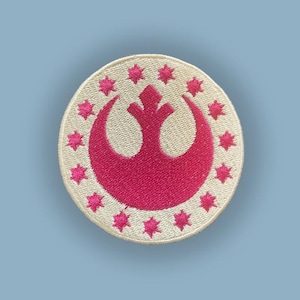 Puede incluir: Parche bordado circular con un símbolo rosa con diseño de media luna y estrellas, rodeado de estrellas rosas sobre un fondo crema. El parche tiene un borde cosido. El fondo es azul claro.