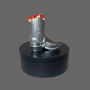May include: A silver cowboy boot match holder filled with red-tipped matches. The boot is sitting on a black circular base.