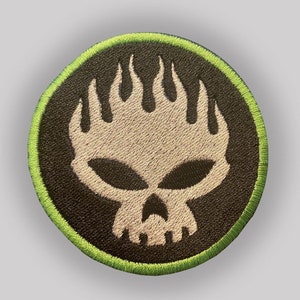 Puede incluir: Un parche bordado en blanco y negro con una calavera y llamas. La calavera es blanca con ojos negros y una boca negra. Las llamas son blancas y están detrás de la calavera. El parche está rodeado por un borde verde.