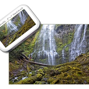 Può includere: Un puzzle da viaggio da 5" x 7" con una cascata su rocce coperte di muschio. Il puzzle è in una scatola con il testo "Tiny Tin Puzzles". Il puzzle completato mostra un paesaggio verde con una cascata. Misura circa 12,7 cm x 17,8 cm.