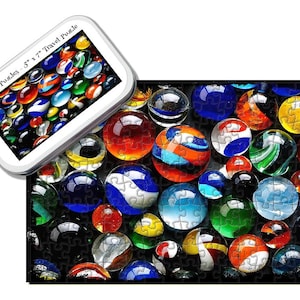 May include: A colorful puzzle featuring a close-up of a variety of marbles in shades of red, blue, green, and white. The marbles are arranged in a random pattern on a black background. The puzzle is made of wood and is 5" x 7". "Tiny Tin Puzzles - 5" x 7" Travel Puzzle" is printed on the tin.