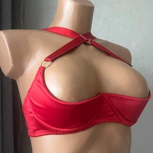 Op de afbeelding: Een rode satijnen bh met een halterband en beugelcups. De bh heeft een uniek ontwerp met een band die over de borst kruist en aan de achterkant vastzit. De bh wordt op een mannequin getoond.
