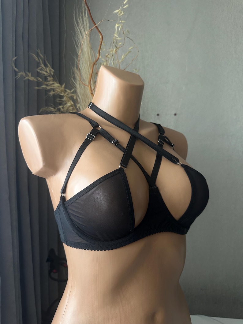 May include: Black lingerie set featuring a bra with a unique harness design. The bra has sheer panels and adjustable straps. The lingerie is displayed on a mannequin against a neutral background.