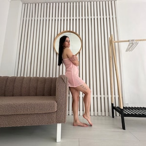 May include: A light pink, spaghetti strap dress. The dress is knee-length and has a textured, ribbed design. The image also shows a brown sofa, a round mirror, and a wooden clothing rack.
