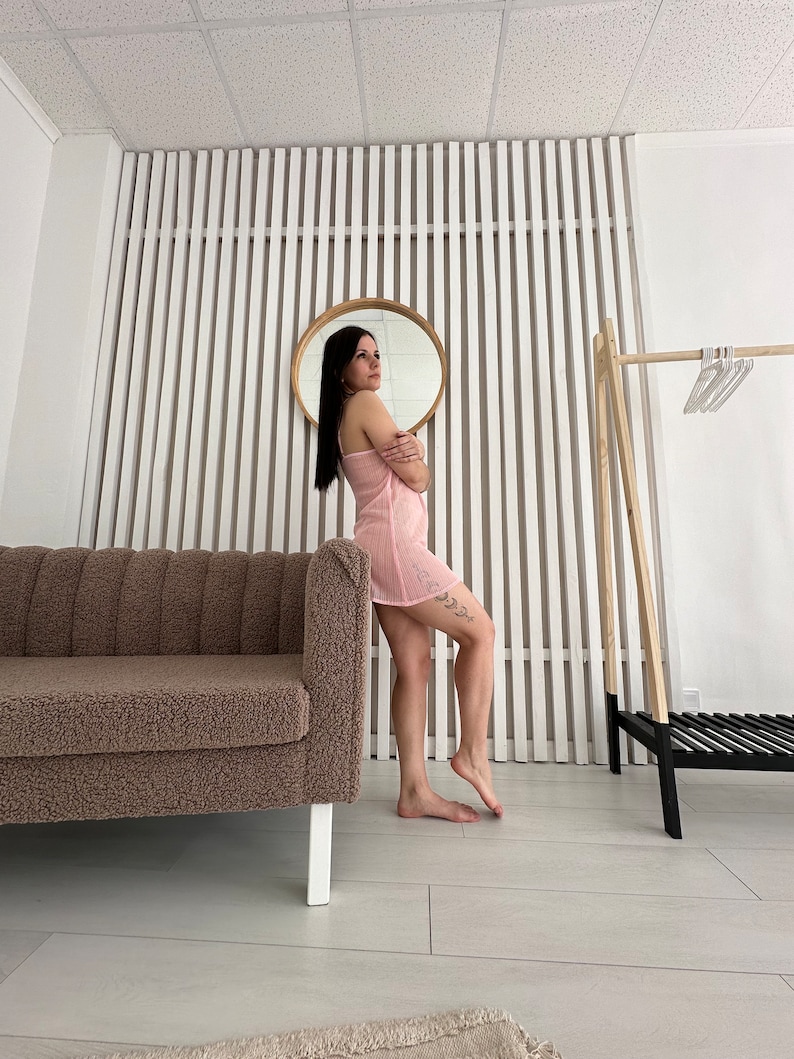 May include: A light pink, spaghetti strap dress. The dress is knee-length and has a textured, ribbed design. The image also shows a brown sofa, a round mirror, and a wooden clothing rack.