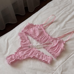 May include: A light pink lingerie set, including a ruffled bralette and matching thong. A pearl necklace is draped across the set. The set is made of a soft, textured fabric. The lingerie is displayed on a white bed.