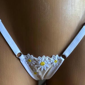Puede incluir: Una tanga blanca con un panel frontal de malla beige transparente decorado con flores de margarita blancas bordadas con centros amarillos. La prenda incluye tiras laterales elásticas blancas finas unidas por anillos de metal plateado.