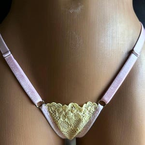 Könnte beinhalten: Ein Tanga mit einem hellgelben floralen Spitzen-Vorderteil und dünnen, verstellbaren hellrosa elastischen Trägern. Das Kleidungsstück wird auf einer Schaufensterpuppe präsentiert und hebt den Kontrast zwischen der zarten Spitzenstruktur und der glatten, satinartigen Oberfläche der Träger hervor.