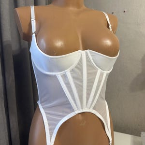 May include: White lingerie set featuring a structured corset with boning and adjustable straps. The set includes a high-cut bottom with garter straps. The sheer fabric and delicate design create a romantic and elegant look.
