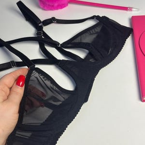 May include: Black lingerie set featuring a sheer mesh design. The bra has adjustable straps and a delicate lace trim. The image also includes a pink pen and a fluffy pink heart-shaped object.