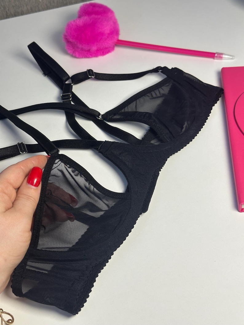 May include: Black lingerie set featuring a sheer mesh design. The bra has adjustable straps and a delicate lace trim. The image also includes a pink pen and a fluffy pink heart-shaped object.