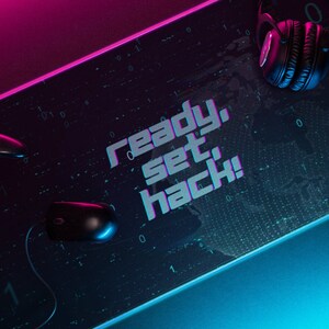 May include: A black gaming mouse, a black video game controller, and black headphones are on a black gaming mat with a blue and pink glow. The mat has a digital pattern and the text "ready, set, hack!" in white.