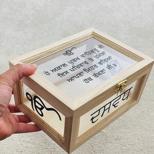 May include: A wooden box with a clear glass top and sides, featuring gold trim and a gold clasp. The box has text in a foreign language on the glass and the side.