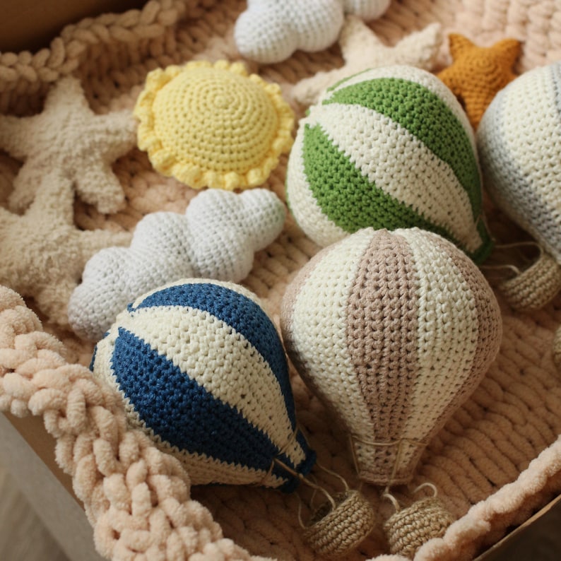May include: A set of crocheted toys including a blue and white striped hot air balloon, a brown and white striped hot air balloon, a green and white striped hot air balloon, a yellow sun, a white cloud, and a white starfish.