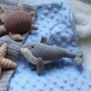 May include: A grey crocheted whale toy with a white belly, a brown crocheted turtle toy, and a beige crocheted starfish toy on a blue textured blanket.