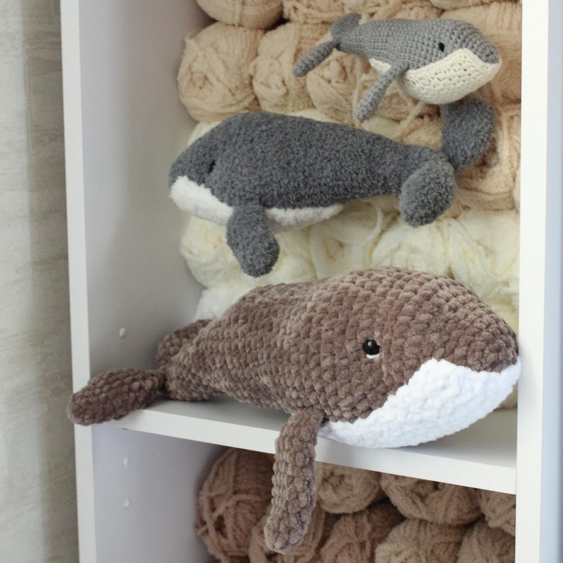May include: Three crocheted whale plushies in different colours and sizes. The whales are sitting on a white shelf with beige yarn balls in the background.