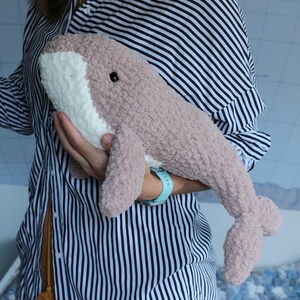 May include: A plush pink and white whale toy with black eyes. The whale is being held by a person wearing a striped shirt.