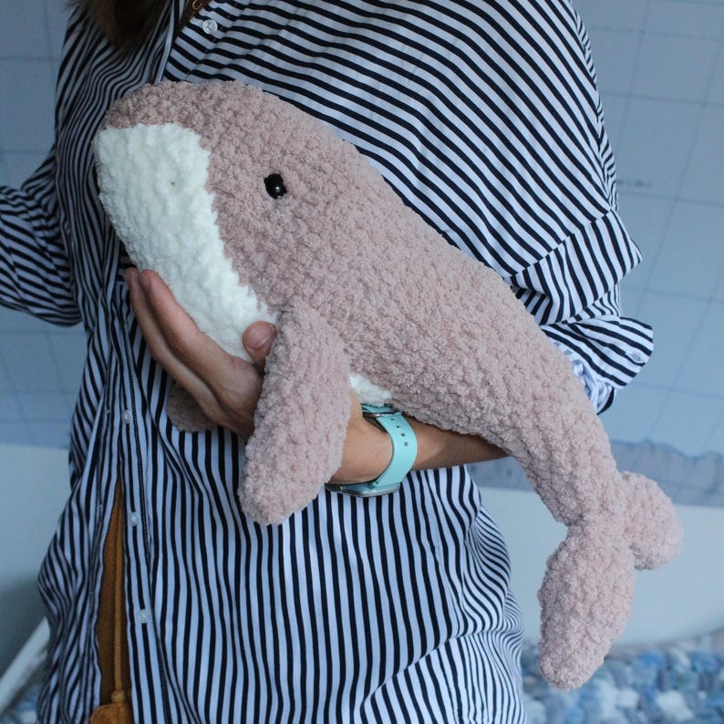 May include: A plush pink and white whale toy with black eyes. The whale is being held by a person wearing a striped shirt.