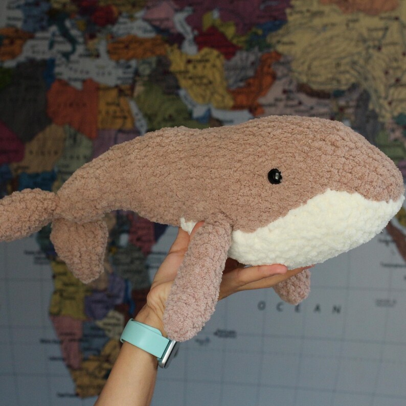 May include: A plush brown whale toy with a white belly and black eyes. The whale is being held in a hand with a light blue watch.