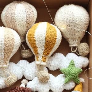 May include: A set of crocheted hot air balloons in various colors and patterns, including white, beige, yellow, and green. The balloons are hanging from white crocheted clouds with a green star.