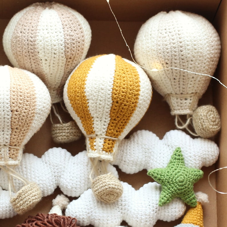 May include: A set of crocheted hot air balloons in various colors and patterns, including white, beige, yellow, and green. The balloons are hanging from white crocheted clouds with a green star.