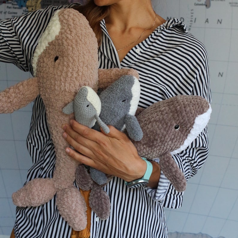 May include: A woman is holding four crocheted whale plushies. The whales are in different sizes and colours, including brown, grey, and white. The largest whale is brown and has a long tail. The smallest whale is white and has a short tail. The two middle whales are grey and have medium-length tails.