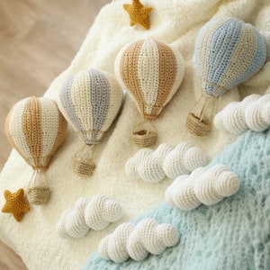 May include: Four crocheted hot air balloons in white, beige, and blue with white crocheted clouds. The balloons are hanging from a white fuzzy surface.