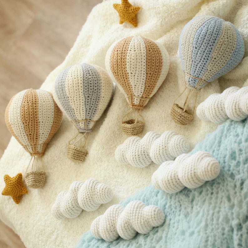 May include: Four crocheted hot air balloons in white, beige, and blue with white crocheted clouds. The balloons are hanging from a white fuzzy surface.