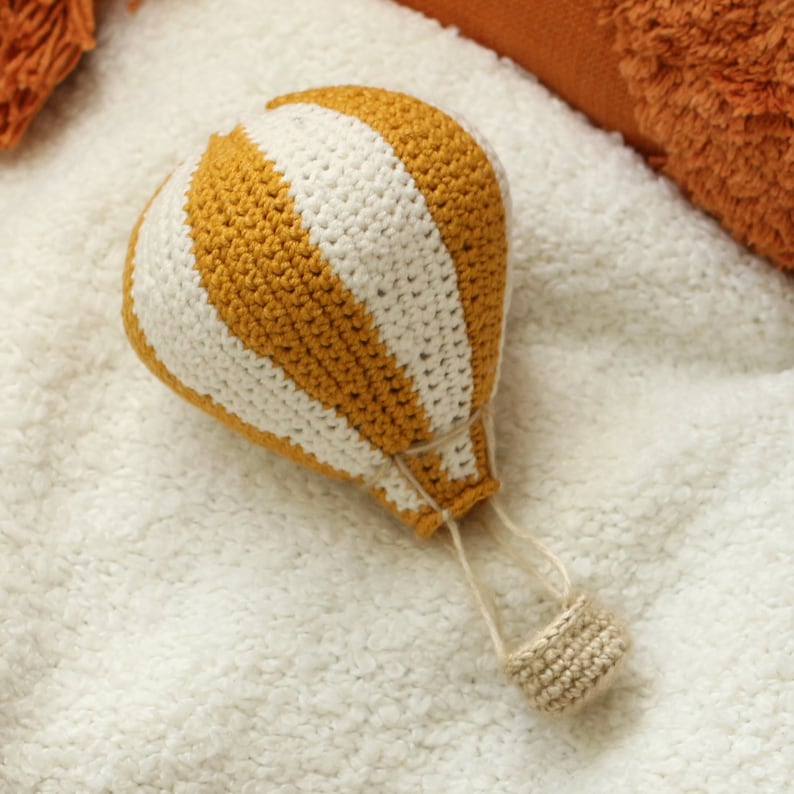 May include: A crocheted hot air balloon toy with yellow and white stripes. The balloon is sitting on a white fluffy surface.