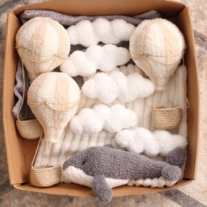 May include: A set of plush toys in a brown box, including three beige hot air balloons, a grey whale, and white fluffy clouds. The balloons have baskets made of woven material.